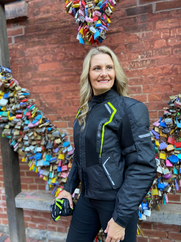 woman standing against a brick wall with a sculpture wearing a gryphon motorcycle jacket smiling
