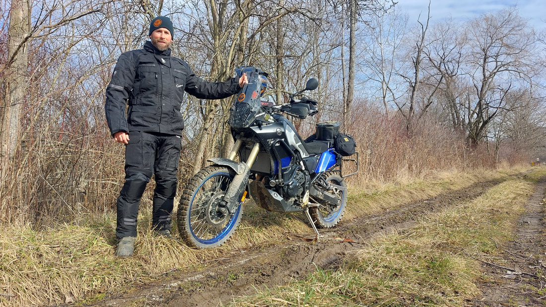 man standing beside his motorcycle wearing gryphon motorcycle jacket and gryphon motorcycle pants on a muddy wooded trail on a cold day. 