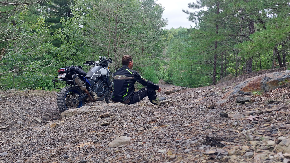 man sitting beside his motorcycle on a rockey forest trail wearing gryphon motorcycle jacket and gryphon motorcycle pants 