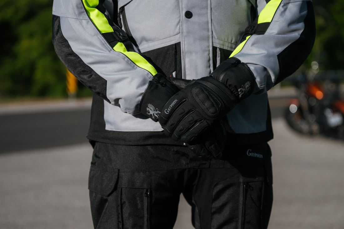 man standing with a motorcycle in the background with his arms crossed in front of him. he is wearing a grey gryphon motorcycle jacket and gloves