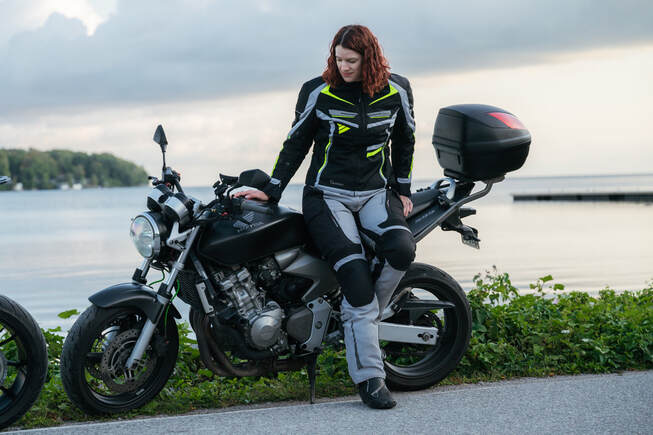 woman leaning against her motorcycle looking downward with a lake in the background wearing gryphon motorcycle gear