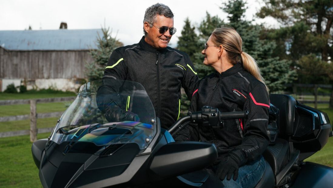 a woman sitting on a motorcycle looking at the man standing beside her. they are smiling at each other. they are both wearing gryphon motorcycle jackets and sunglasses 