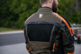 man standing on a road with a motorcycle in the background wearing a gryphon motorcycle jacket showing a large vent panel in the back of the jacket 