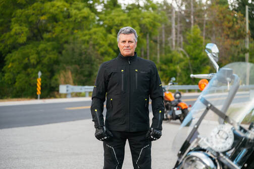man standing on the road with a motorcycle in the background and foreground wearing gryphon protective motorcycle jacket, gloves and pants