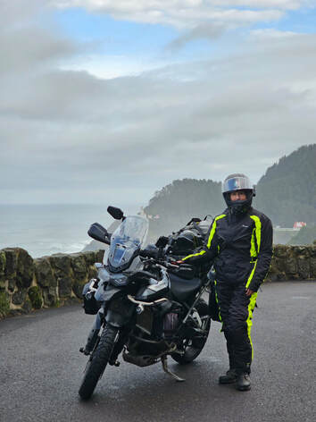 woman standing beside her motorcycle on a rainy day wearing gryphon rain gear with the ocean and mountains in the background