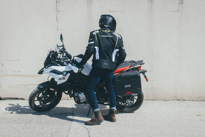man facing away beside his motorcycle against a concrete wall wearing jeans and a gryphon motorcycle jacket with a helmet 
