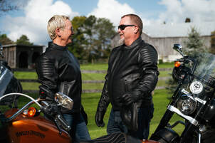 a man and a woman standing behind their motorcycles, looking at each other, smiling. they are wearing sunglasses and gryphon leather motorcycle jackets and gloves