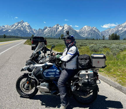 a man sitting on his motorcycle wearing gryphon protective motorcycle gear at the side of the road with a meadow and snow covered mountains in the background