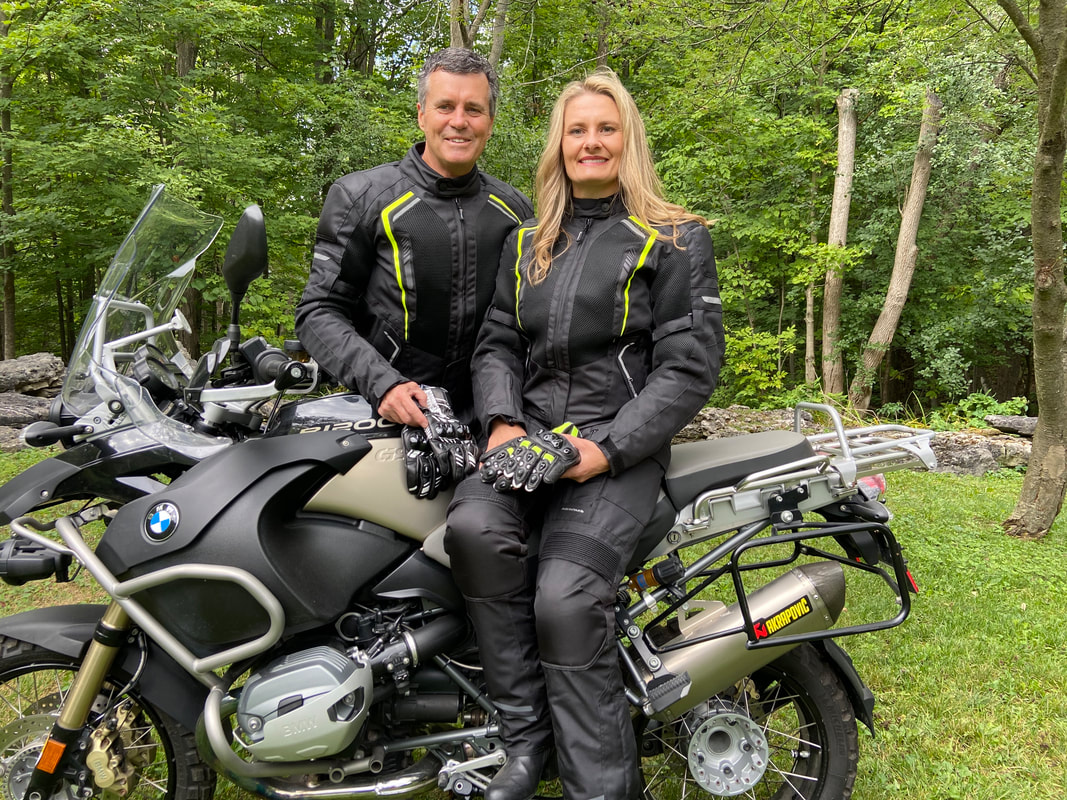 woman sitting on a motorcycle with a man standing behind wearing Gryphon motorcycle gear. both people are smiling. 