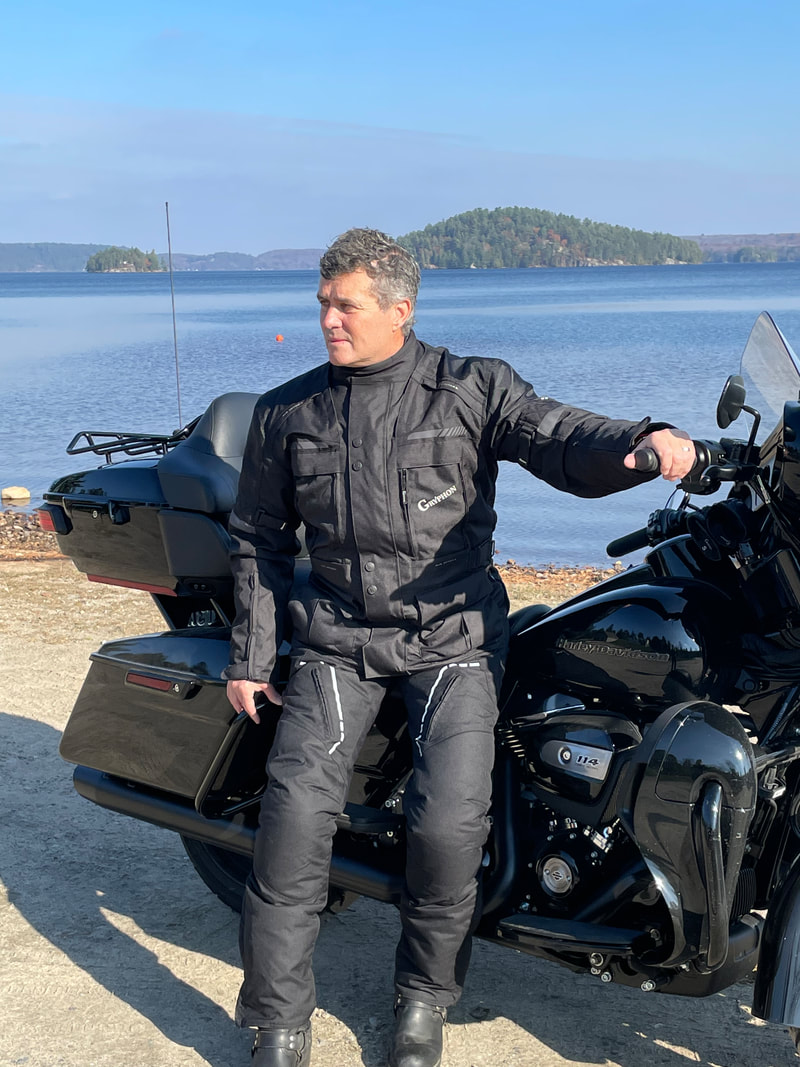 man sitting on a harley motorcycle at the beach wearing a black gryphon motorcycle jacket and black gryphon motorcycle pants