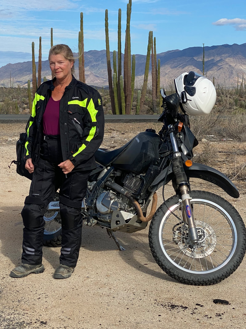woman standing beside a motorcycle in the desert with cacti behind, wearing a gryphon motorcycle jacket and gryphon motorcycle pants 