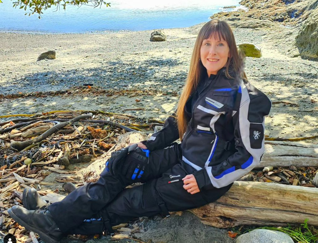 woman sitting on a log at the beach wearing gryphon motorcycle gear smiling